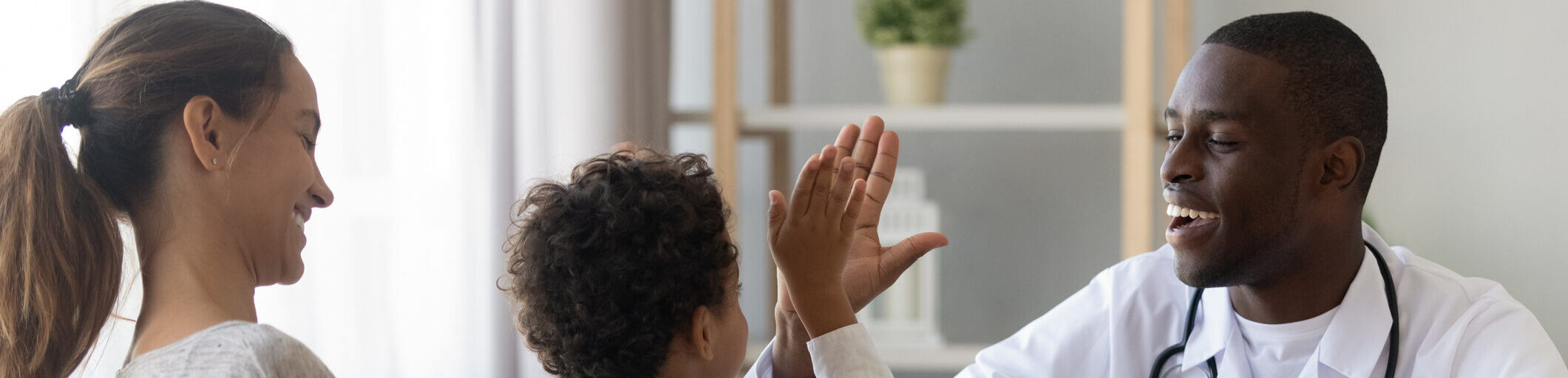 Smiling pediatric doctor giving a high-five to a young child during a checkup while the child's parent watches, representing compassionate, family-focused healthcare.