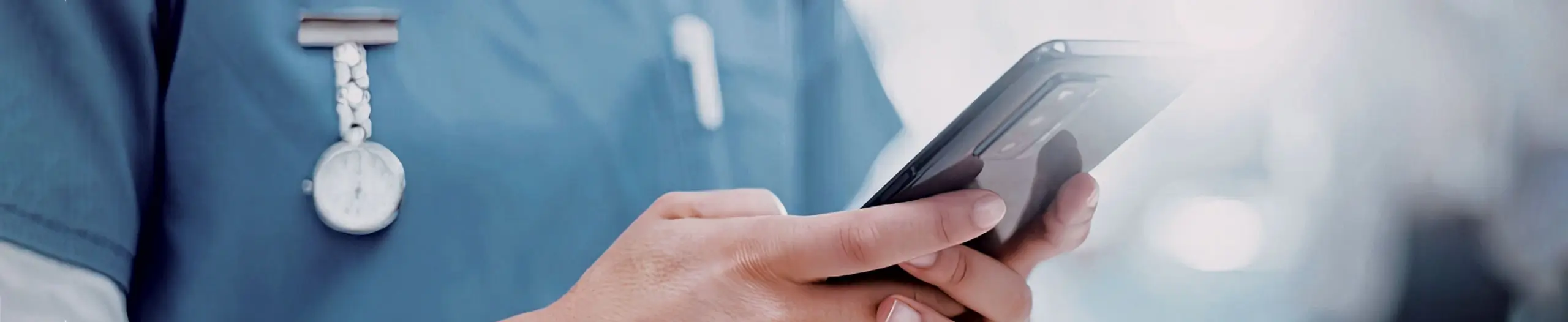 Healthcare professional in blue scrubs using a smartphone with a stethoscope around their neck.