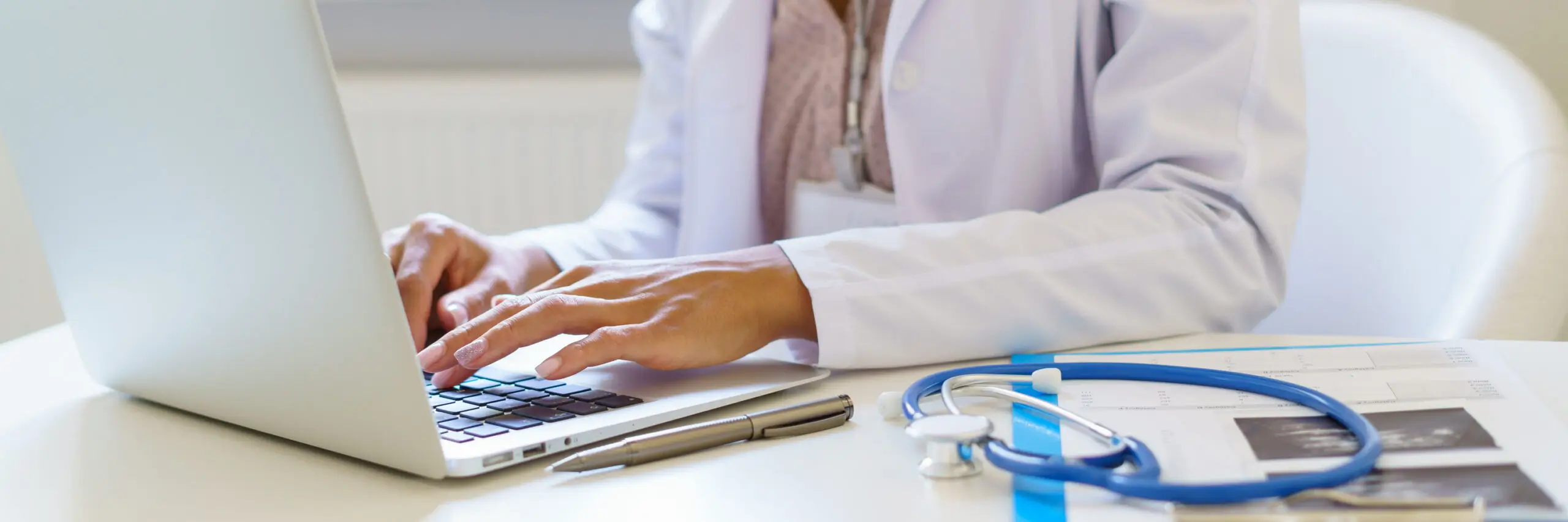 Healthcare professional in a white lab coat typing on a laptop at a medical desk with a stethoscope, patient records, and pen, representing digital healthcare and medical administration.
