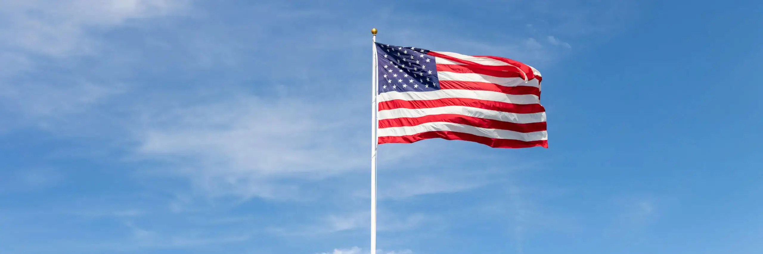 United States flag flying on a white flagpole under a clear blue sky with soft clouds in the background.