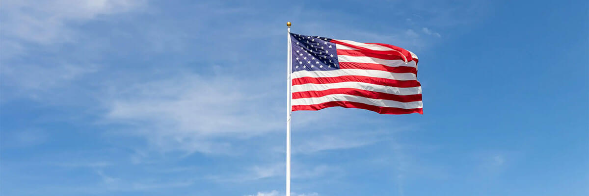 United States flag flying on a white flagpole under a clear blue sky with soft clouds in the background.