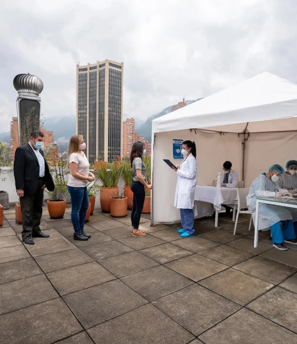 People wearing face masks stand in line at an outdoor COVID-19 testing station while healthcare workers assist patients under a white medical tent.