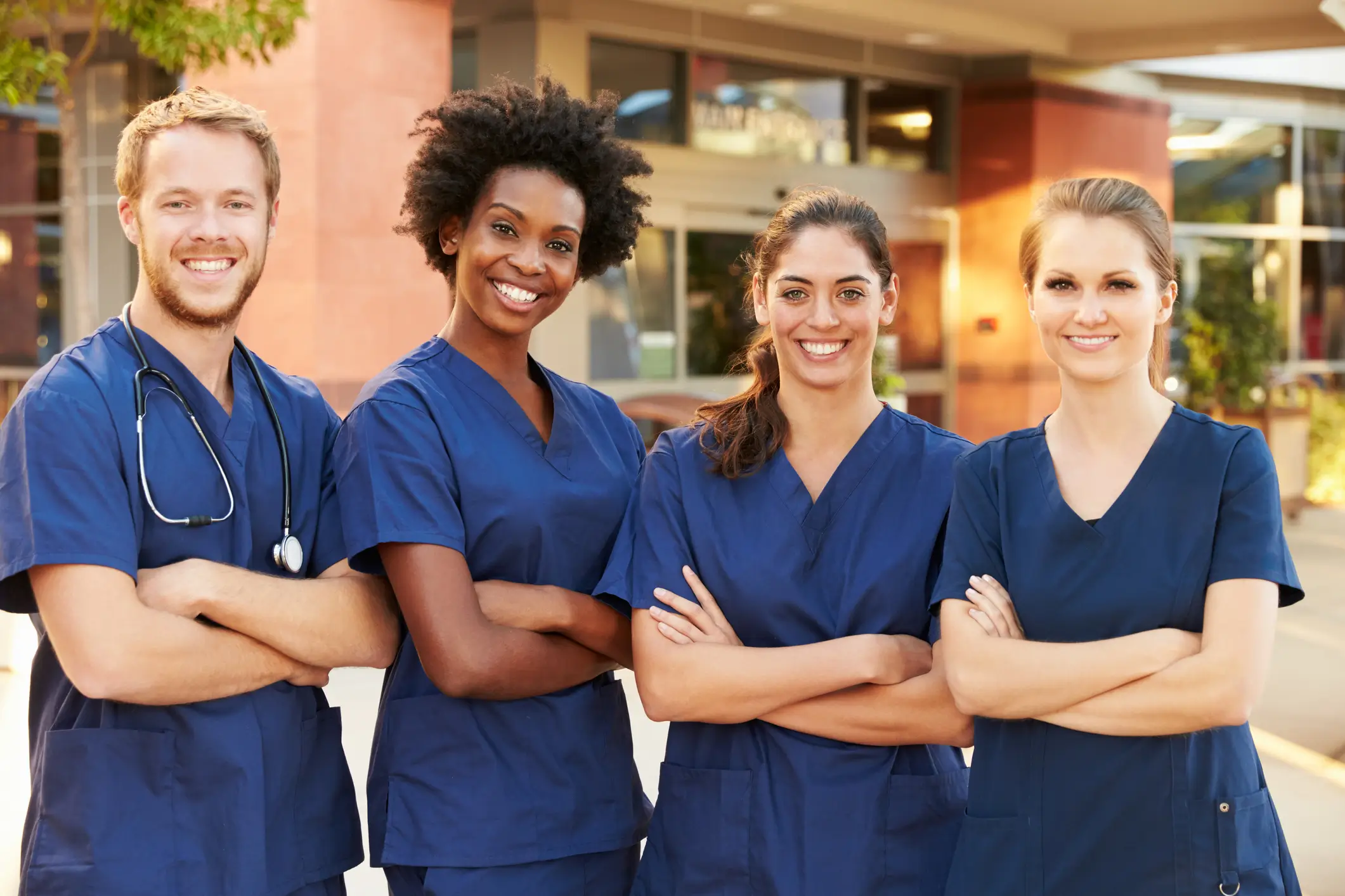 Four smiling healthcare professionals in blue scrubs stand together outside a medical facility, representing a diverse team of nurses and medical staff.