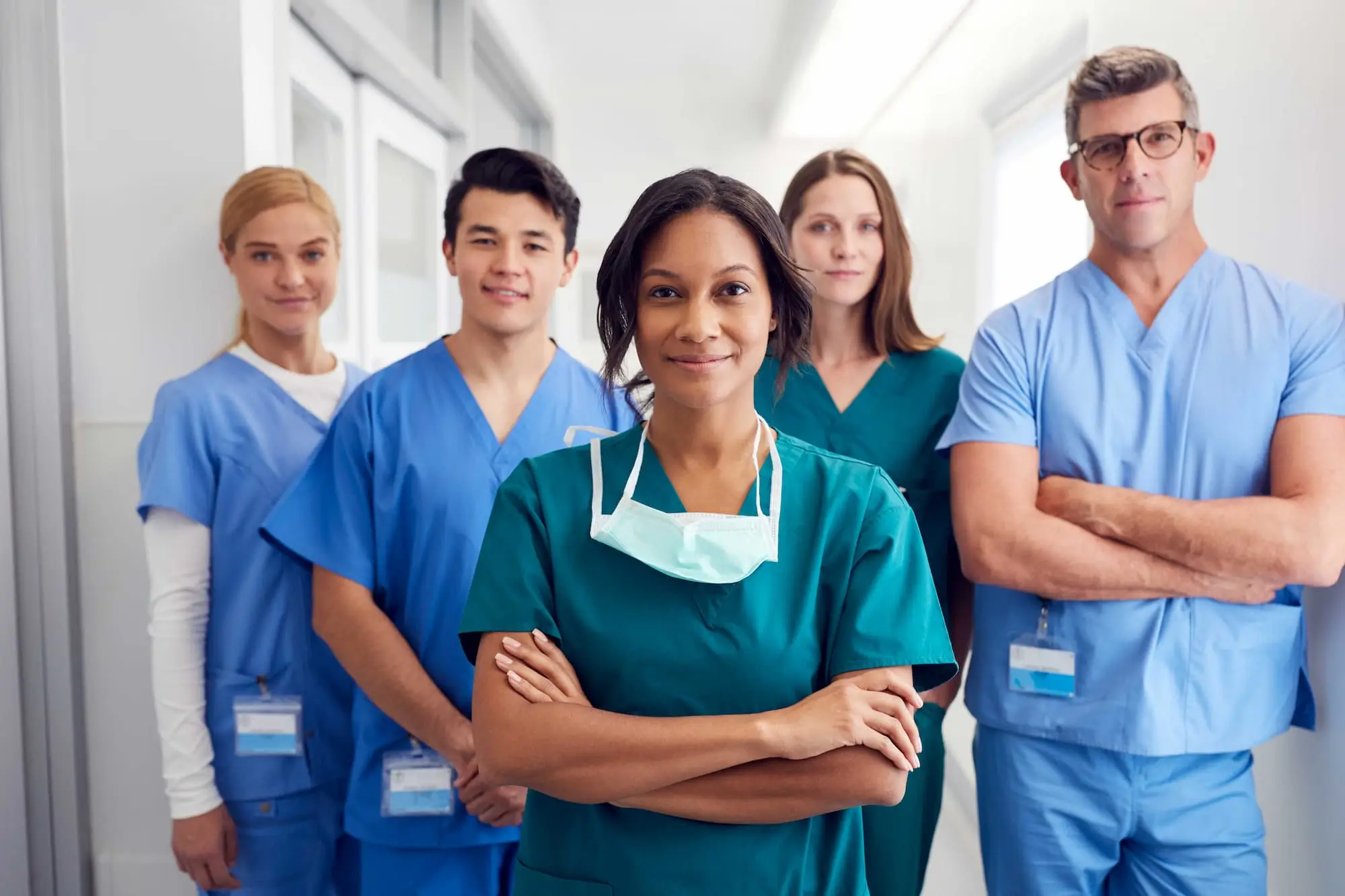 A diverse team of healthcare professionals wearing medical scrubs stands together in a hospital hallway, with a nurse in the foreground and colleagues behind her.