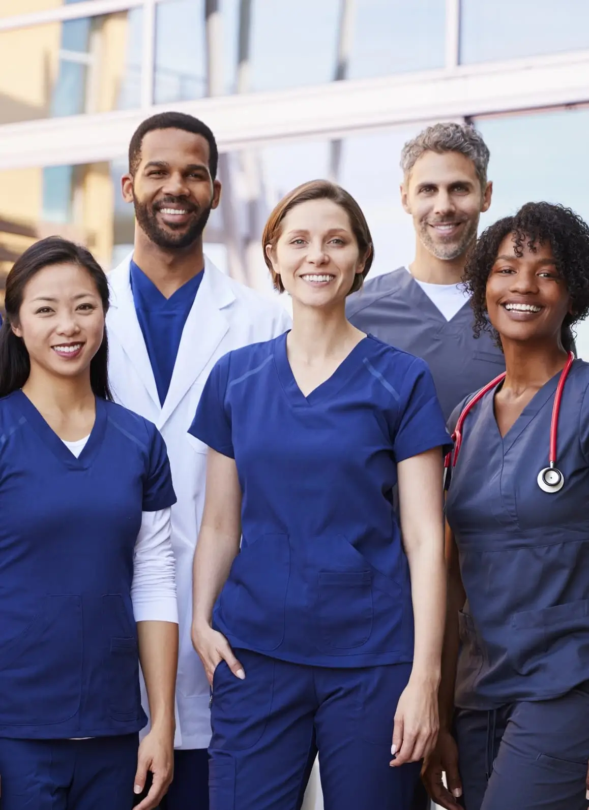 A diverse group of smiling healthcare professionals, including doctors and nurses in scrubs and lab coats, standing together outside a modern medical facility.