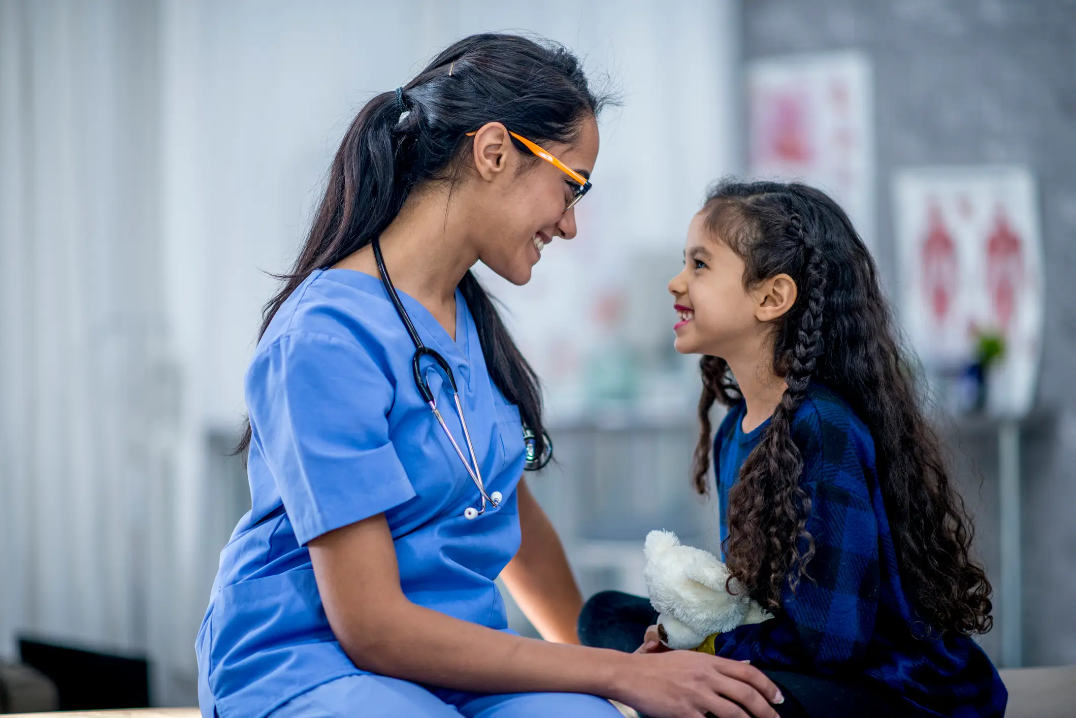 Smiling pediatric nurse in blue scrubs talking with a young girl holding a stuffed toy during a friendly healthcare visit.