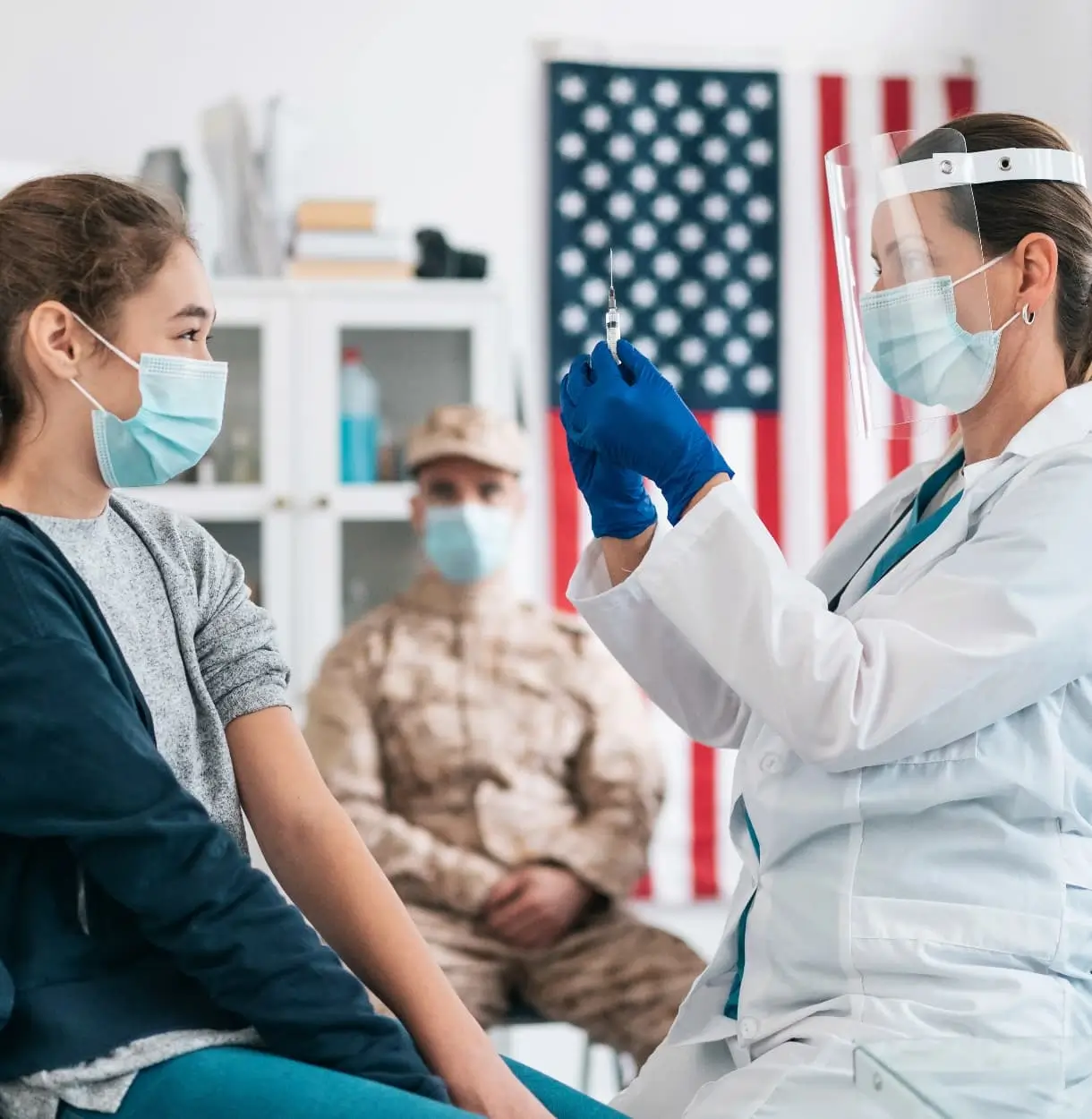 Healthcare professional wearing a face shield and mask preparing a vaccine for a masked teenage patient while a military service member waits in the background.