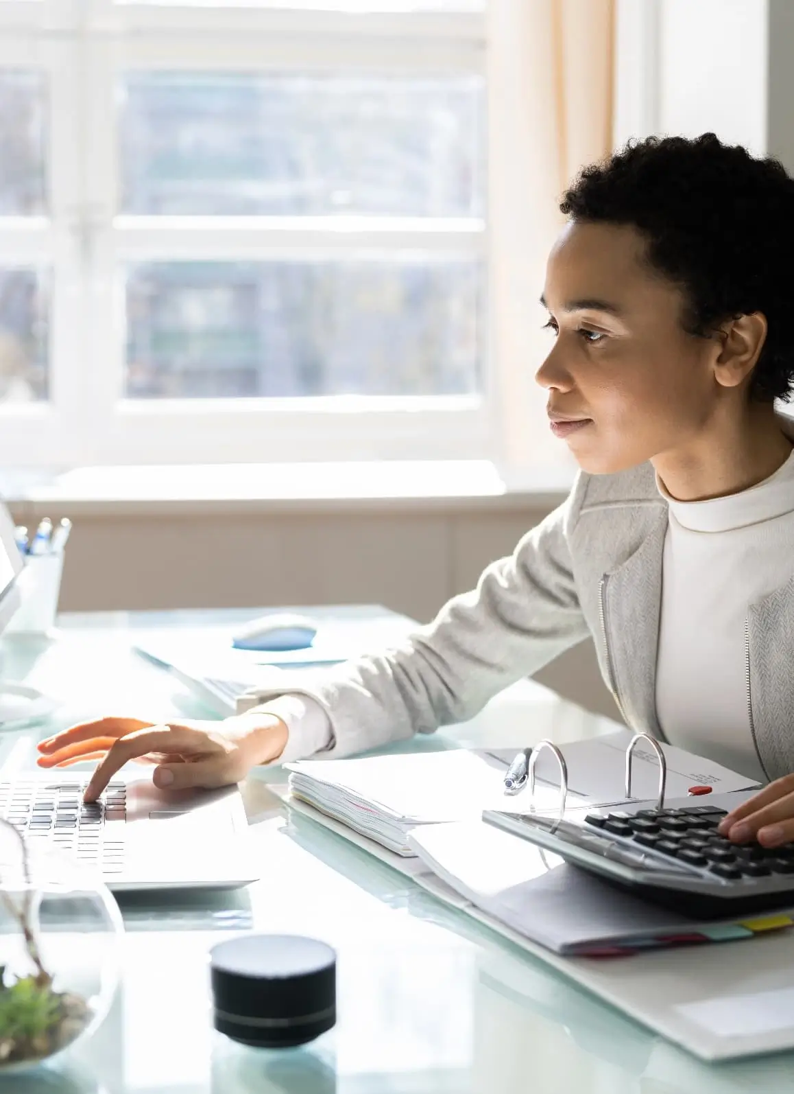 Young healthcare professional working on a laptop at a desk with paperwork, a calculator, and office supplies in a bright workspace.